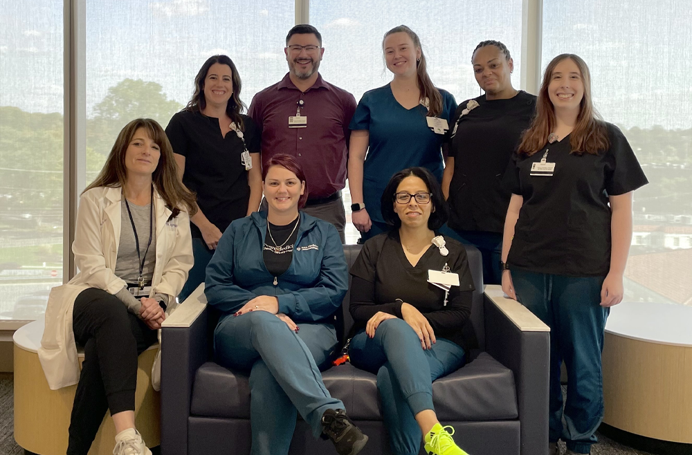 A group of healthcare professionals in scrubs and lab coats pose together in a brightly lit room with large windows.